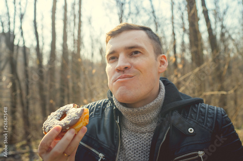 Man enjoying doughnut in forest From below of happy man in warm jacket eating glazed doughnut standing in sunny woods