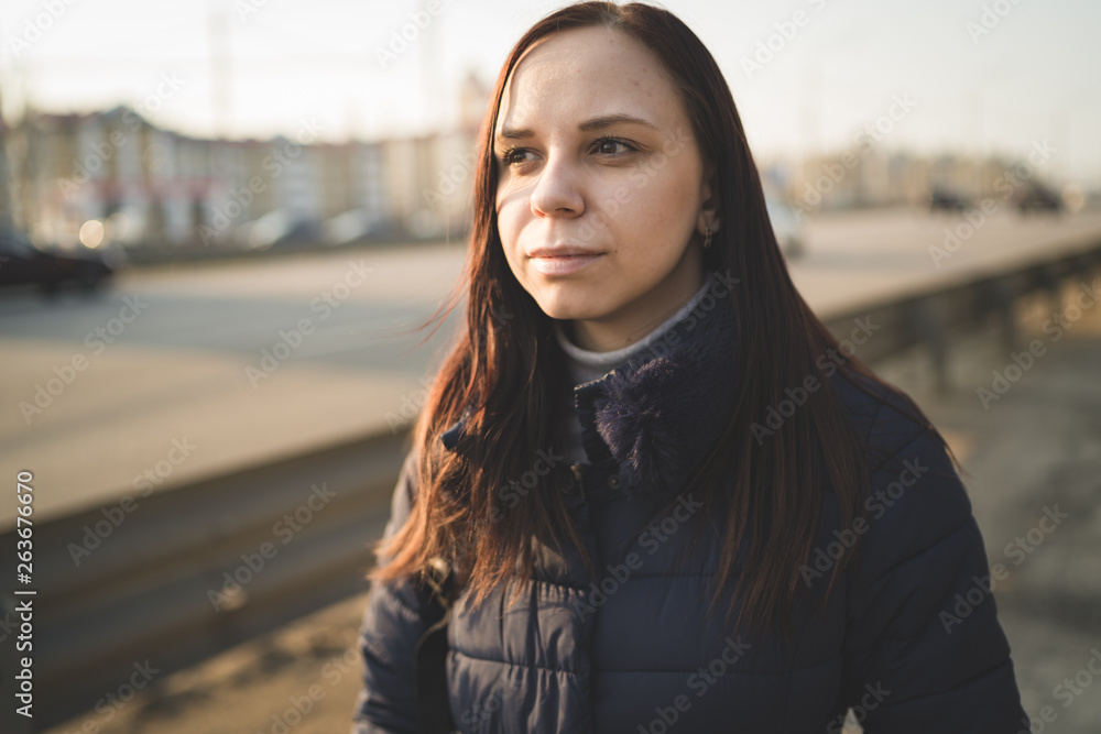 Young woman on city roadside Casual brunette in warm jacket standing on pavement of city roadway looking pensively away