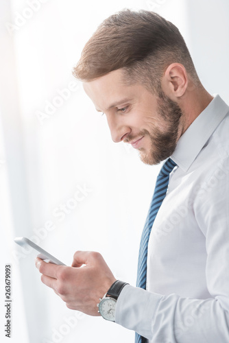 cheerful bearded man in suit smiling while using smartphone