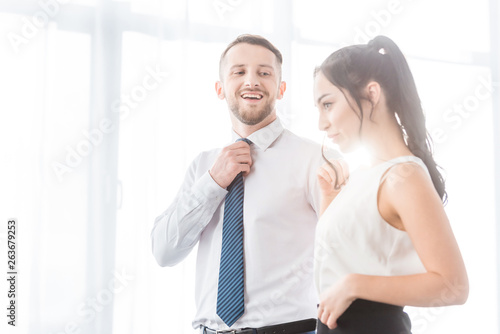 cheerful man touching tie and looking at attractive woman