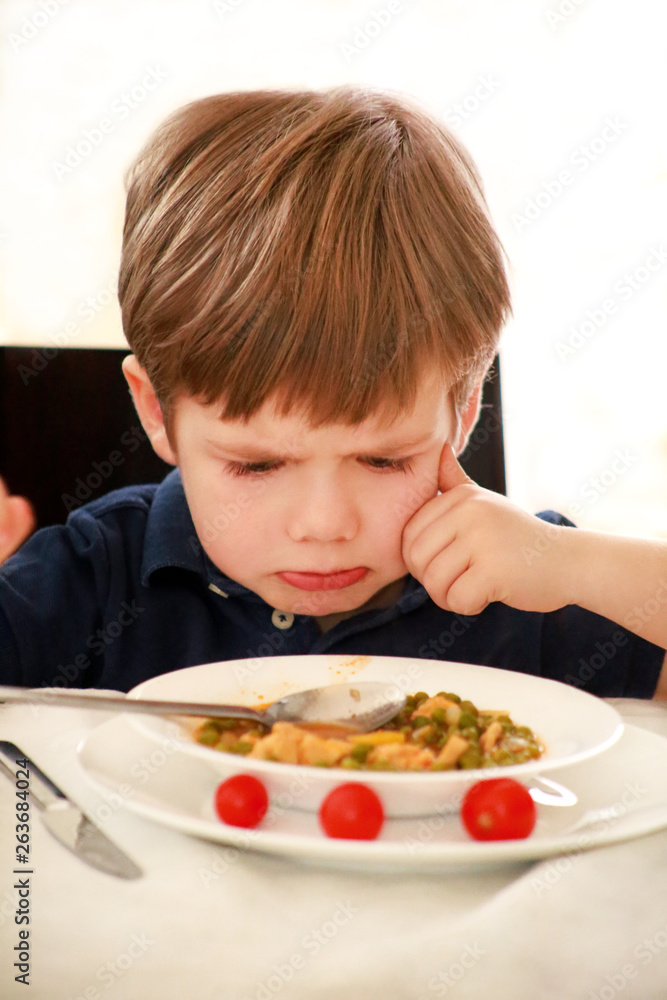 Hungry child sitting in chair at table in kitchen and despises, disapproves of hot meal, because he does not like lunch. Kid is having of cooked peas with tomato for lunch. Healthy diet food concept. Hungry child sitting in chair at table in kitchen and despises, disapproves of hot meal, because he does not like lunch. Kid is having of cooked peas with tomato for lunch. Healthy diet food concept.