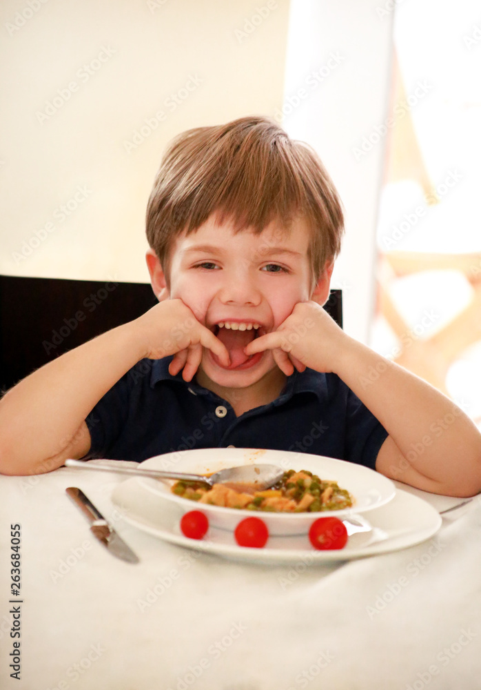 Hungry child sitting in chair at table in kitchen and eating with spoon of cooked peas with tomato. Kids meal and healthy diet food concept. Happy and cute little boy enjoying in good lunch at home.