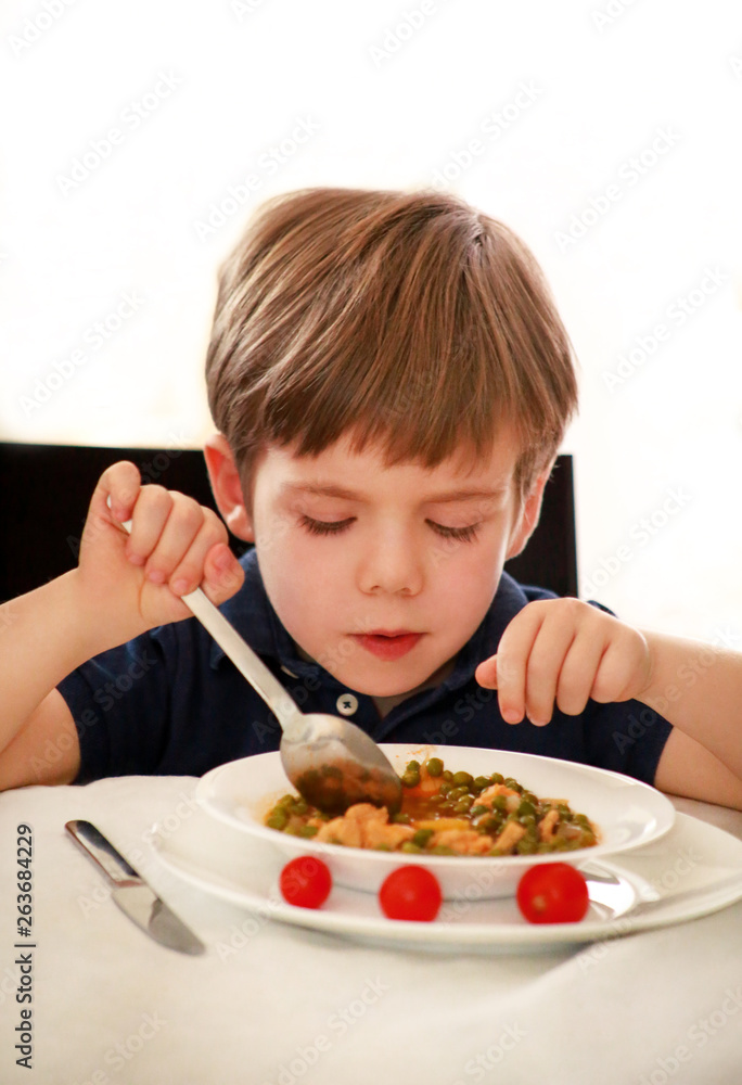 Hungry child sitting in chair at table in kitchen and eating with spoon of cooked peas with tomato. Kids meal and healthy diet food concept. Happy and cute little boy enjoying in good lunch at home.