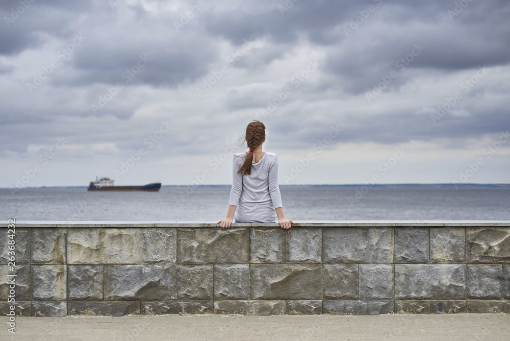 Rear view of a girl sitting on the embankment of the river. A ship is sailing on the horizon. There are heavy clouds in the sky. Rear view of a girl sitting on the embankment of the river. A ship is sailing on the horizon. There are heavy clouds in the sky.
