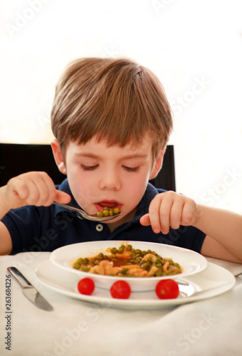 Hungry child sitting in chair at table in kitchen and eating with spoon of cooked peas with tomato. Kids meal and healthy diet food concept. Happy and cute little boy enjoying in good lunch at home.