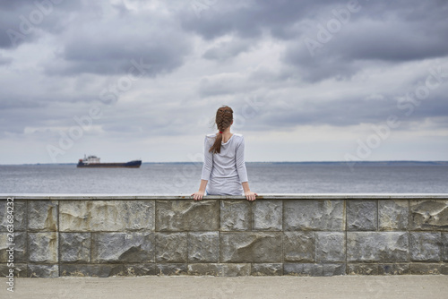 Rear view of a girl sitting on the embankment of the river. A ship is sailing on the horizon. There are heavy clouds in the sky.