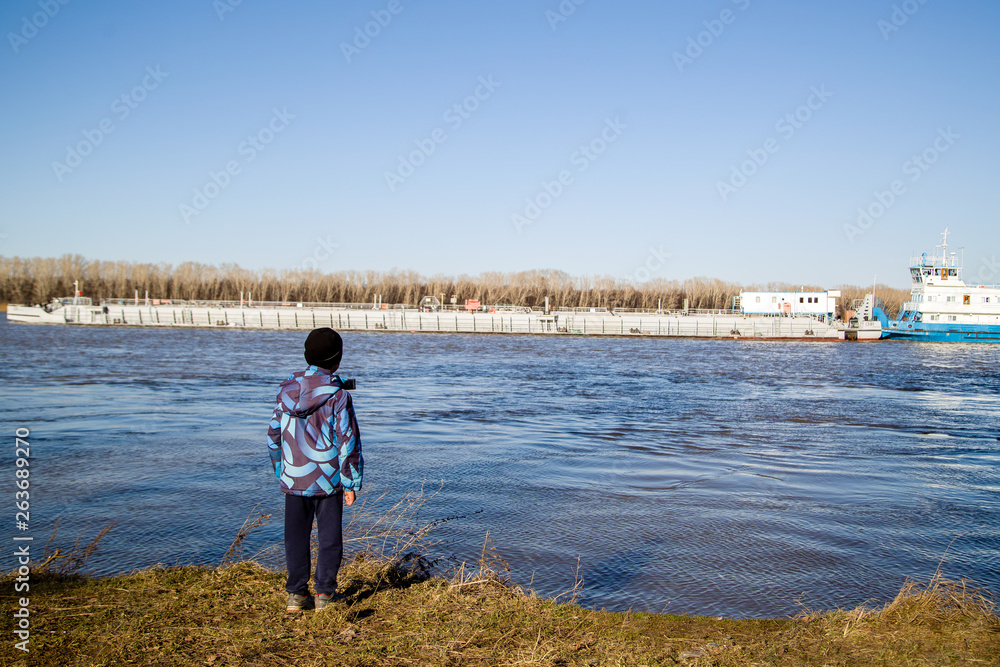 The boy on the shore looking at the tug with a barge