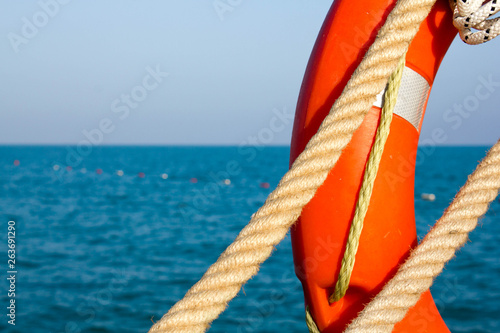 Orange lifeline and sea ropes on the background of the sea and blue sky. Marine ropes and life preserver hanging on a green post on the background of blue sea and sky. Sea ropes. Close up.