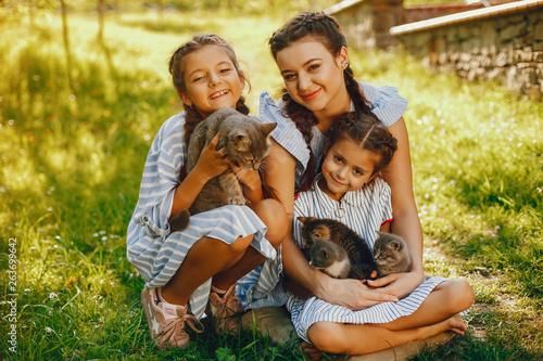 three beautiful and cute girls in blue dresses with beautiful hairstyles and make-up sitting in a sunny green garden and playing with a cats