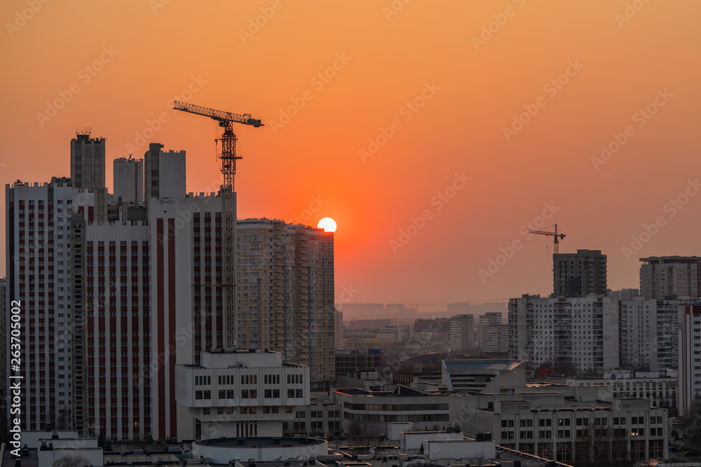 Sunset sun hiding behind city's high-rise buildings, orange cloudless sky cityscape