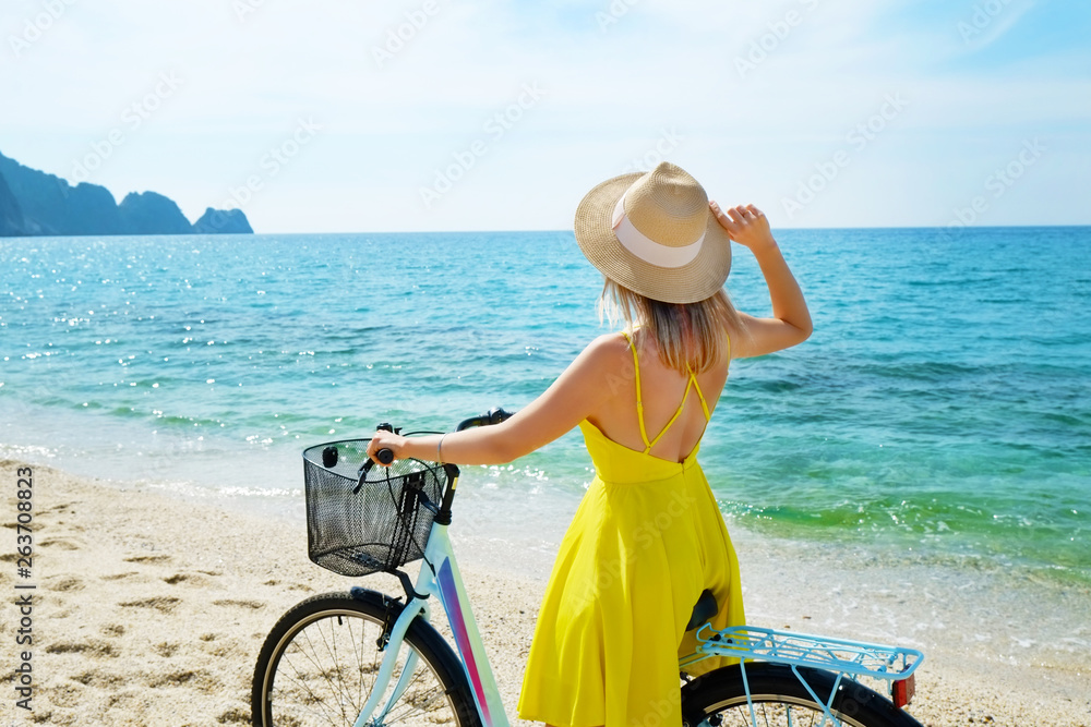 Young carefree woman in bright yellow dress with bicycle at ocean beach. Unrecognizable female wearing broad brim straw hat biking on sandy sea shore on sunny day. Copy space, background.