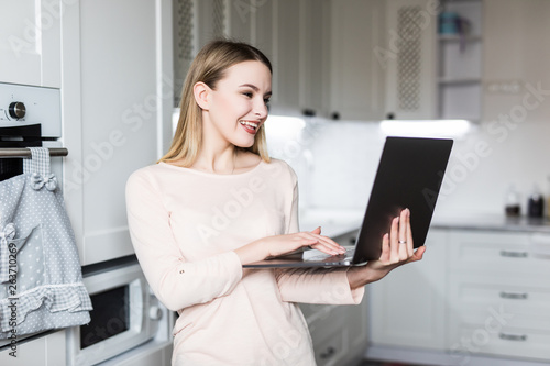 Happy female looking at laptop while standing in kitchen in morning.