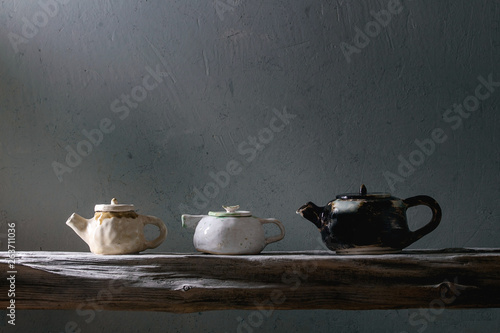 Variety of craft handmade ceramic teapots for tea ceremony standing on old wooden shelf in dark room.