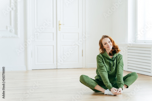 Photo of delighted redhead female sits on lotus pose, wears tracksuit, being in good body shape, has curly ginger hair, poses on floor, copy space for your promotional content. Sport concept