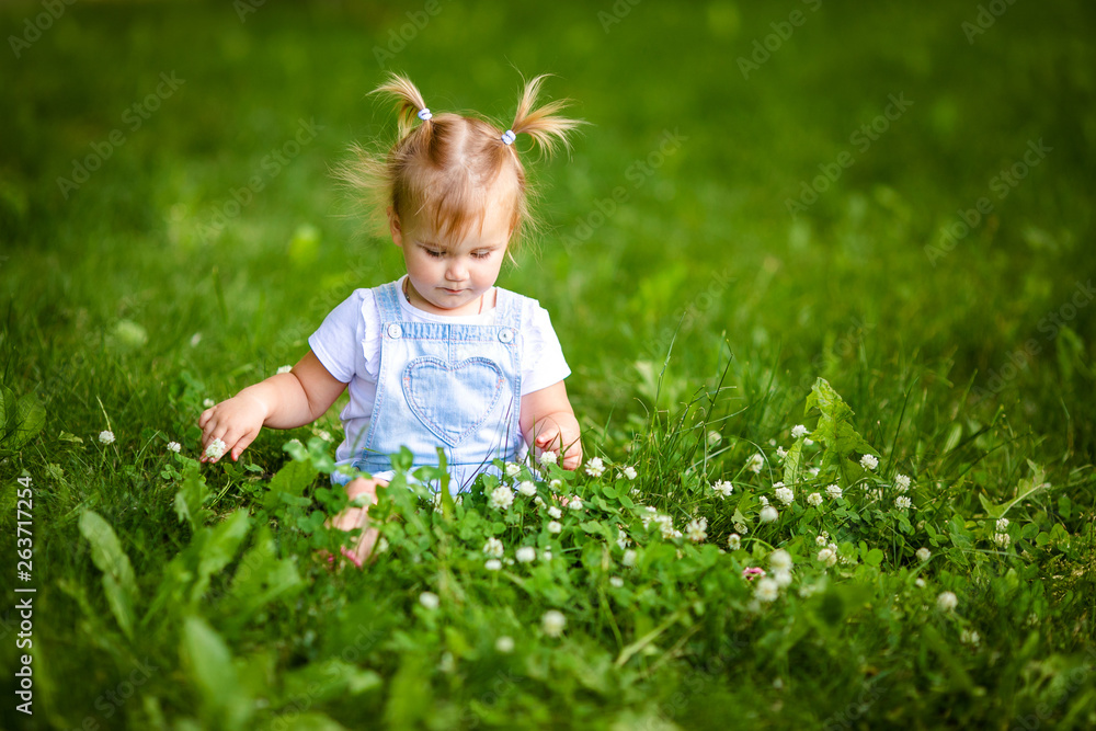 Portrait of adorable little blonde girl lying on grass and propping up her face at summer green park. Baby girl with white flowers Portrait of adorable little blonde girl lying on grass and propping up her face at summer green park. Baby girl with white flowers