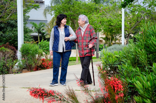 Cheerful female carer and an older woman with a walking stick stroll in a city park. Both are in a great mood.
