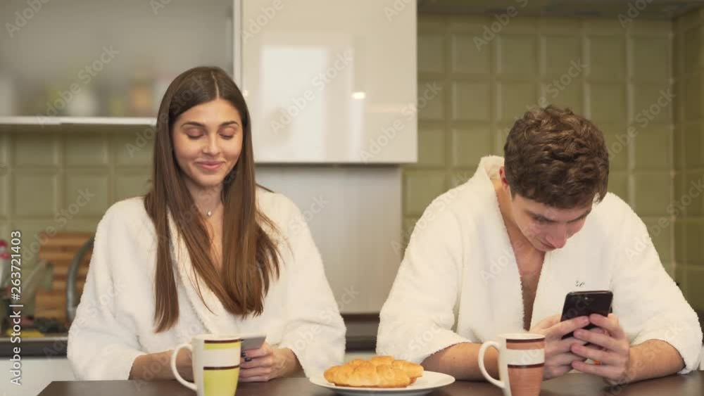 Funny couple in a white bathrobes are sitting in a kitchen and clicking in a phones. Woman is smiling and keeking in on man's phone and he is smiling and hiding it. Funny couple in a white bathrobes are sitting in a kitchen and clicking in a phones. Woman is smiling and keeking in on man's phone and he is smiling and hiding it.