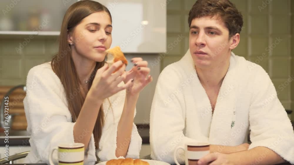 Funny couple in a white bathrobes are sitting in a kitchen. Woman is eating a bun, man is trying to bite it, but woman is hiding the bun and smiling. Couple is in love and kissing. Funny couple in a white bathrobes are sitting in a kitchen. Woman is eating a bun, man is trying to bite it, but woman is hiding the bun and smiling. Couple is in love and kissing.