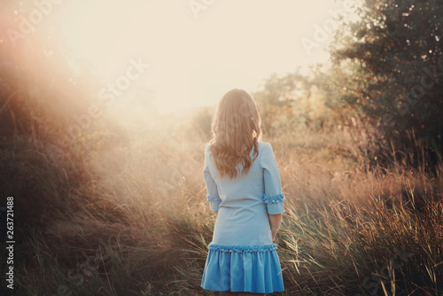 Back view of a beautiful young woman or girl on very green meadow watching the sunset enjoying nature summer evening outdoors. Sunshine.