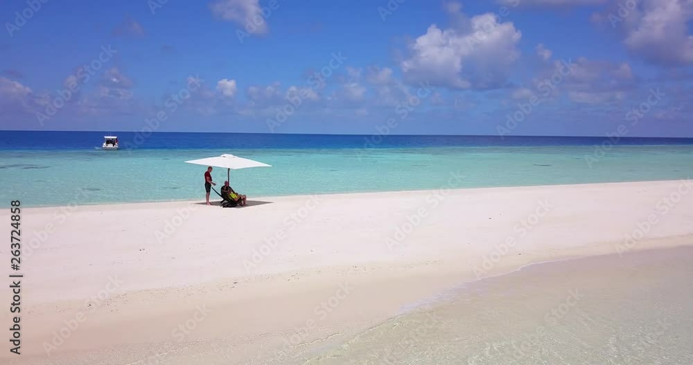 Three friends are enjoying the turquoise Caribbean sea from a distance under a white parasol. Wide-angle drone footage, vertical zoom in a slide Three friends are enjoying the turquoise Caribbean sea from a distance under a white parasol. Wide-angle drone footage, vertical zoom in a slide
