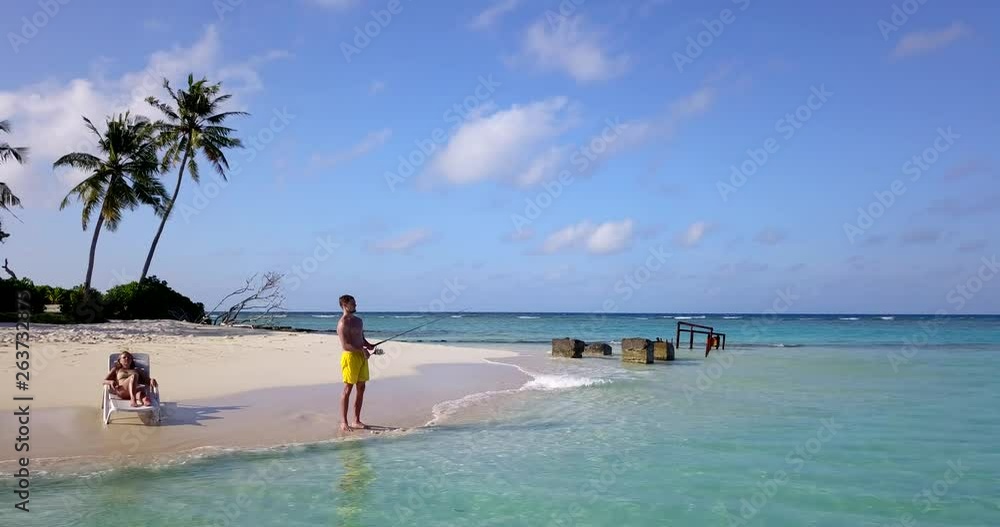 Man fishes on beach while woman sunbathes on tropical island, aerial push in 4k Man fishes on beach while woman sunbathes on tropical island, aerial push in 4k