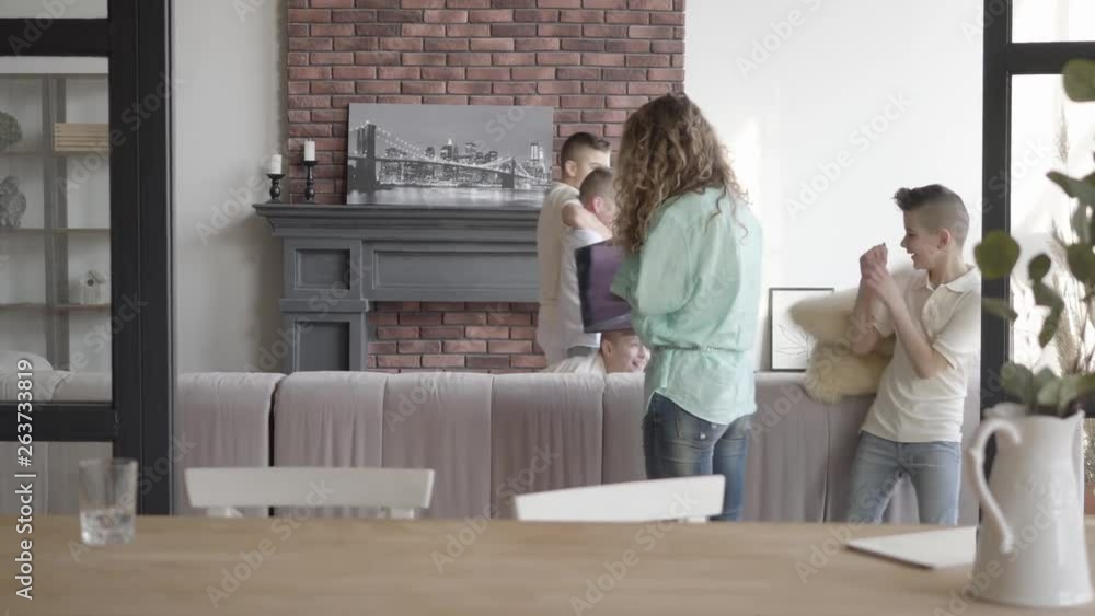 Young mother holding laptop trying to calm her four teen sons running araund and playing with each other. Happy big noizy family. Vase with plant in the foreground. Camera moves left Young mother holding laptop trying to calm her four teen sons running araund and playing with each other. Happy big noizy family. Vase with plant in the foreground. Camera moves left