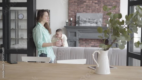 Tired young mother trying to calm herself standing near the wall with glass of water while her teen sons running araund playing with each other in the background. Big noizy family photo