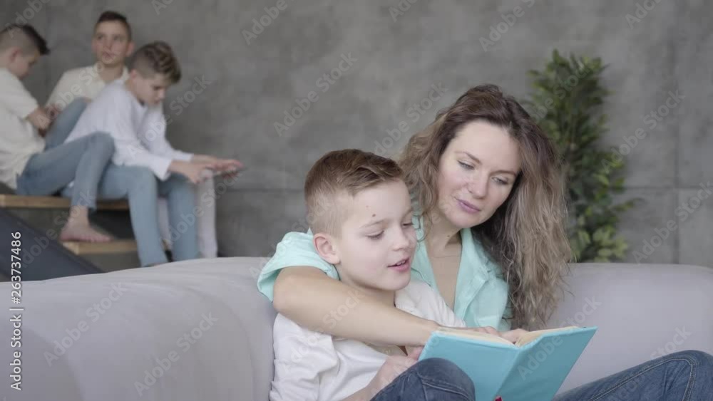 Portrait of pretty young mother reading book to her son while the rest of her teen kids playing with each other in the background sitting on stairs with tablet. Big friendly noizy family Portrait of pretty young mother reading book to her son while the rest of her teen kids playing with each other in the background sitting on stairs with tablet. Big friendly noizy family