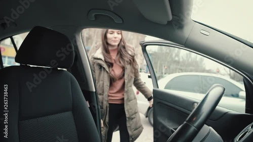 A young girl in a long khaki-colored jacket with long hair, a brunette, gets behind the wheel of her car and starts driving out by turning on the reverse gear. photo