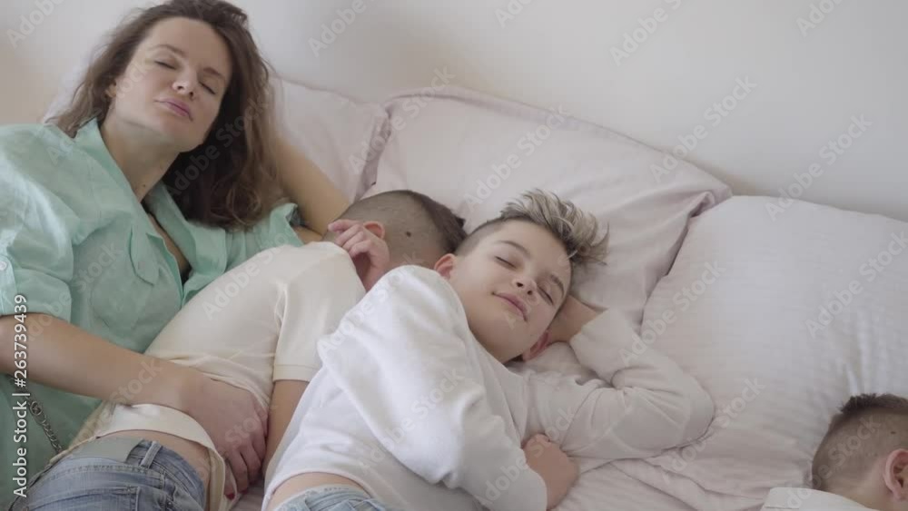 Portrait young mother with her teen sons sleeping together in the big bed. One boy sitting on the chair. Happy loving family concept. Quiet hour in the family with many kids Portrait young mother with her teen sons sleeping together in the big bed. One boy sitting on the chair. Happy loving family concept. Quiet hour in the family with many kids