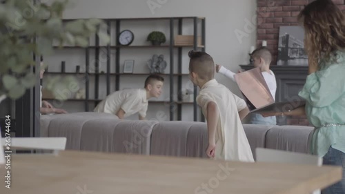 Cute young mother holding laptop trying to talk by cell phone while her four teen sons running araund playing with each other in the background. Happy big noizy family. photo