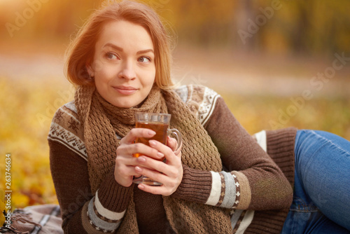 Beautiful young girl sitting on a plaid in the fall leaves in the park, drinking hot tea. Concept of autumn warmth, atmosphere and comfort