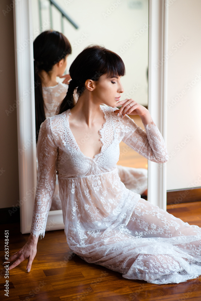 Sensual young beautiful woman in white lace nightgown sitting near the mirror at home. Before the wedding ceremony. Lingerie and nightwear Sensual young beautiful woman in white lace nightgown sitting near the mirror at home. Before the wedding ceremony. Lingerie and nightwear
