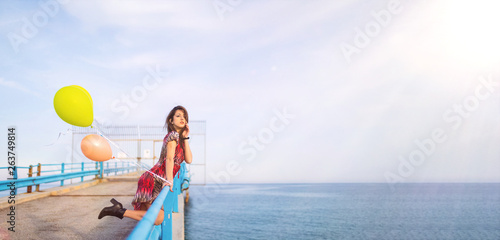 Estate. Ragazza con vestito rosso allegra su un pontile con dei palloncini colorati riempiti di elio vicino la spiaggia sul Mare.