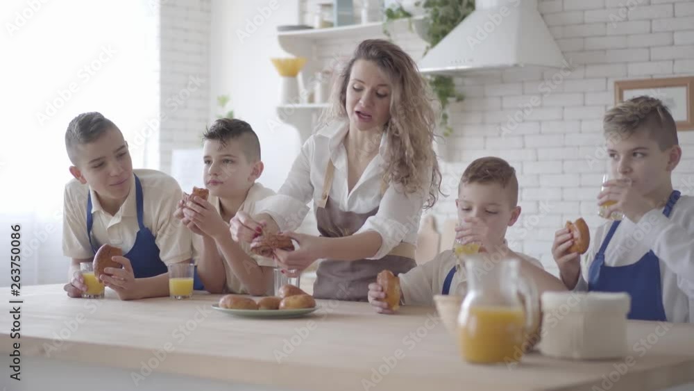 Attractive happy woman and her four teen sons eating pies and drinking orange juice in the kitcken standing at the table. Everybody wearing aprons. Kids helping mother to cook food Attractive happy woman and her four teen sons eating pies and drinking orange juice in the kitcken standing at the table. Everybody wearing aprons. Kids helping mother to cook food