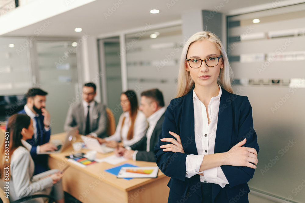 Portrait of attractive blonde businesswoman in formal wear holding crossed arms while standing in board room In background colleagues having meeting. Portrait of attractive blonde businesswoman in formal wear holding crossed arms while standing in board room In background colleagues having meeting.