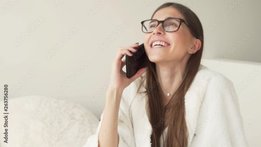 Beautiful young girl is talking on the phone, smiling and laughing. Girl is wearing a glasses and a white bathrobe. Beautiful young girl is talking on the phone, smiling and laughing. Girl is wearing a glasses and a white bathrobe.
