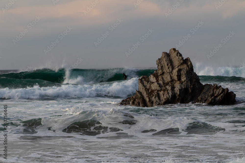 Fototapeta premium a surfer practicing surfing on Atxabiribil beach, next to a large rock, in the photo the sunset begins