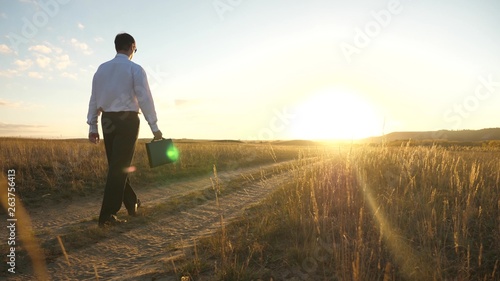 businessman in sunglasses goes down the country road with a briefcase in his hand. The entrepreneur works in a rural area. a farmer inspects land at sunset. agricultural business concept. © zoteva87