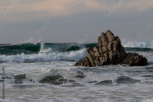 a surfer practicing surfing on Atxabiribil beach, next to a large rock, in the photo the sunset begins