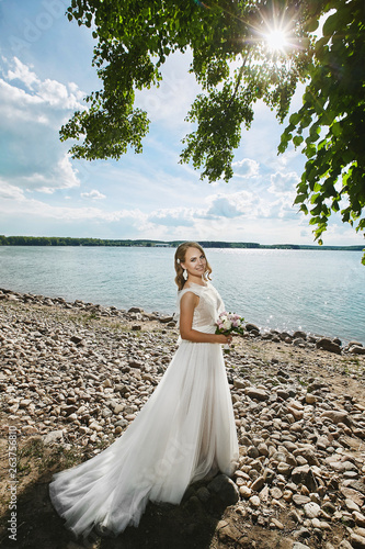 Beautiful young happy bride with wedding hairstyle and with a bouquet of fresh flowers in her hands walking by the coast of the lake