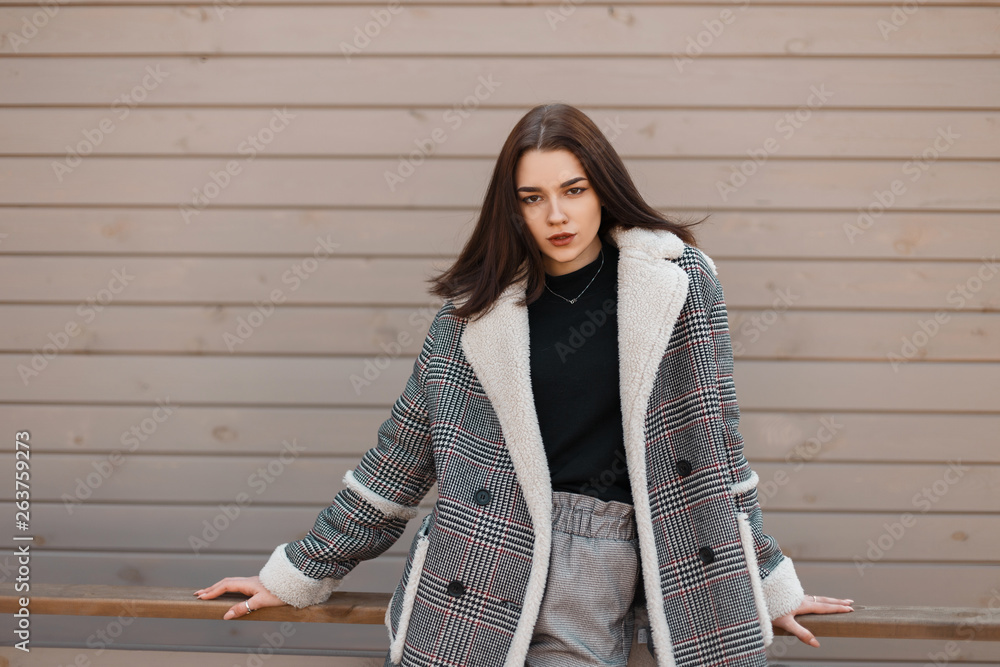 Beautiful modern young woman in trendy black T-shirt in a gray elegant  checked jacket in fashionable vintage pants is standing outdoors near the  wooden wall. European girl fashion model. Youth. Stock Photo