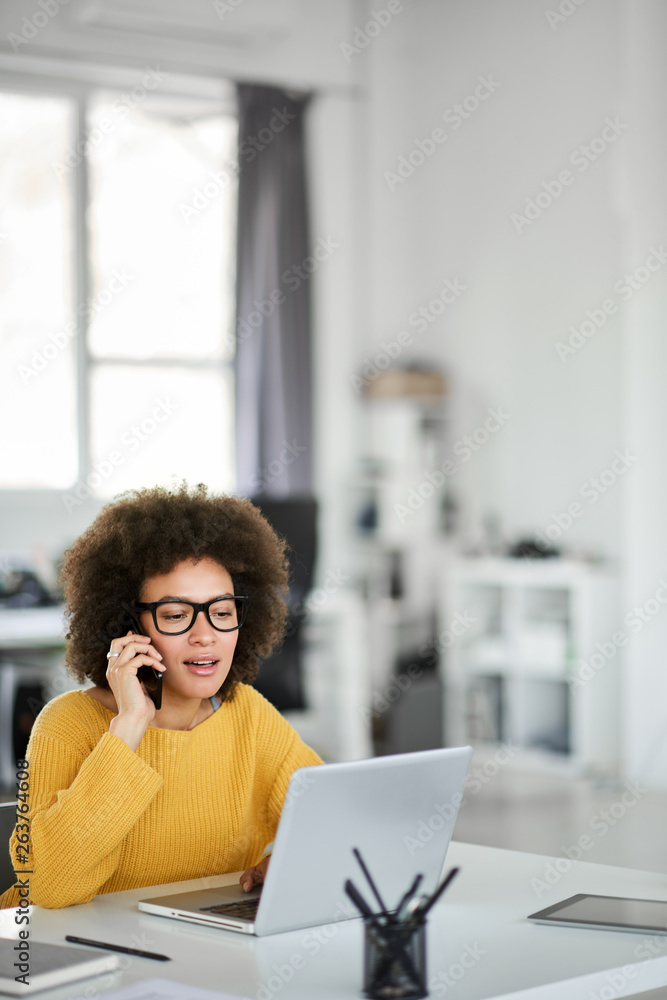 © nenadaksic - Charming mixed race businesswoman dressed casual using smart phone and laptop at the same time while sitting in modern office.