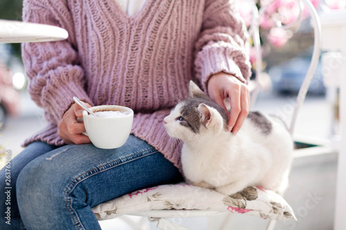 Young woman is drinking coffee and stroking pet cat in spring garden. Girl is sitting outdoors in cafe on blooming city streets with pink magnolia flowers. photo