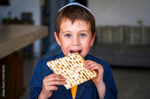 Cute Caucasian child in a yarmulke taking a bite from a traditional Jewish matzo unleavened bread in a room. photo