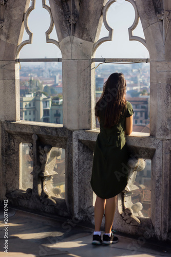 Attractive girl in a green dress and sneakers, with long dark hair, turns her back and looks out the window at the view of the city from the roof of the Milan Cathedral. Milano Duomo, Italy, observe.