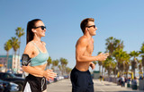 fitness, sport and technology concept - happy couple with earphones and arm bands running over venice beach background in california