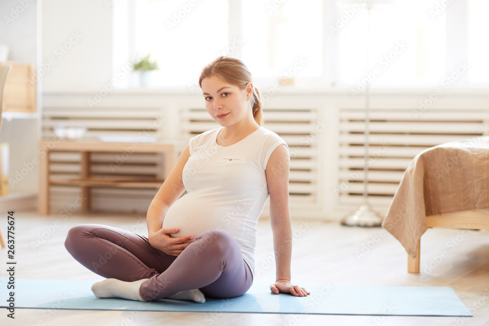 Full length portrait of young pregnant woman sitting on yoga mat while exercising at home and looking at camera, copy space Full length portrait of young pregnant woman sitting on yoga mat while exercising at home and looking at camera, copy space