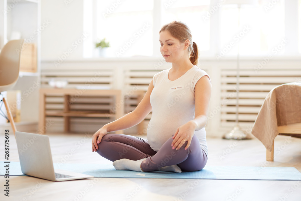 Full length portrait of young pregnant woman sitting on yoga mat watching workout tutorial at home, copy space Full length portrait of young pregnant woman sitting on yoga mat watching workout tutorial at home, copy space