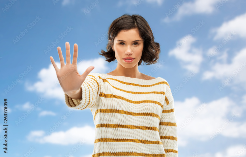 Fototapeta premium restriction, prohibition and people concept - young woman in striped pullover making stopping gesture over blue sky and clouds background